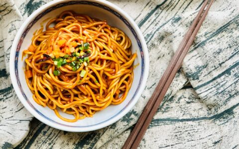 A bowl of traditional Wuhan noodles garnished with vegetables, served with wooden chopsticks.