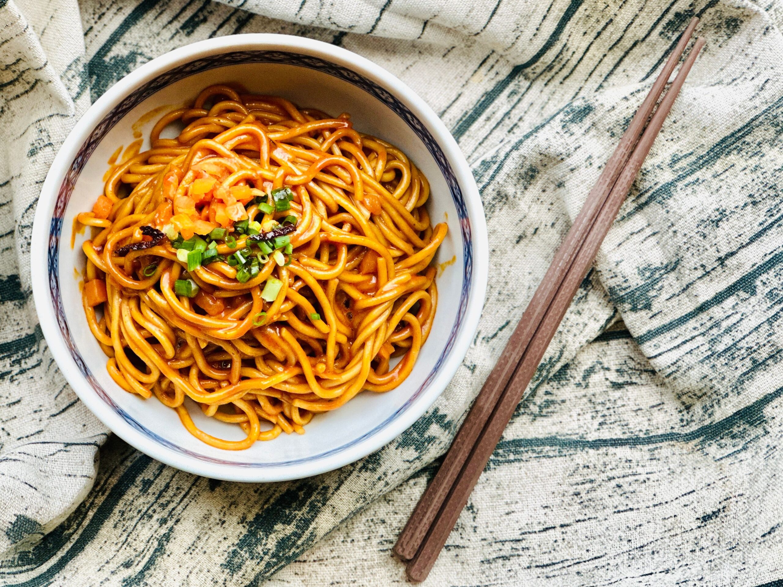 A bowl of traditional Wuhan noodles garnished with vegetables, served with wooden chopsticks.