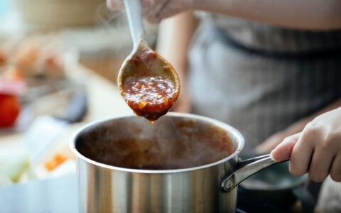 Crop anonymous females in aprons cooking sauce for pasta in saucepan in kitchen together